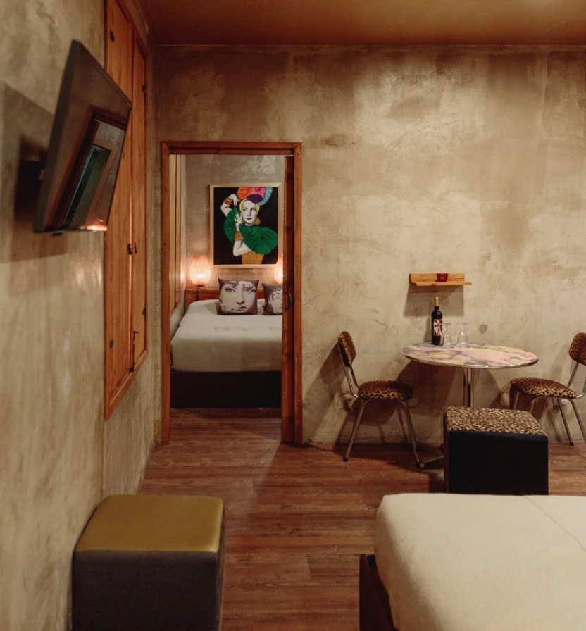 Cozy hotel room corridor with bed, chairs, table, and framed artwork.