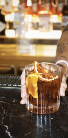 Hand holding a mug of amber cocktail with foam and garnish on bar counter