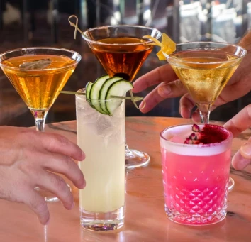 Hands holding three colorful martini cocktails: orange, clear, and pink with ice.