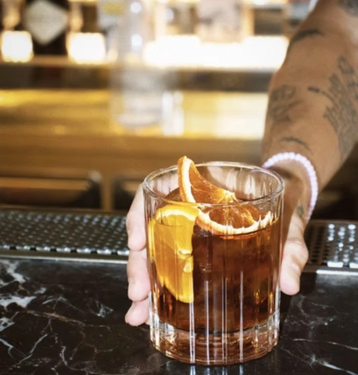 Bartender holds a glass of amber cocktail with an orange peel garnish.