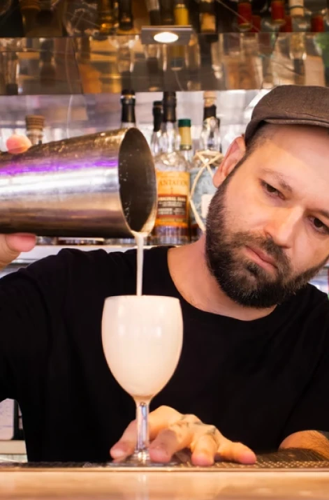 Man pours a drink from a jigger into a glass with bottles behind him.