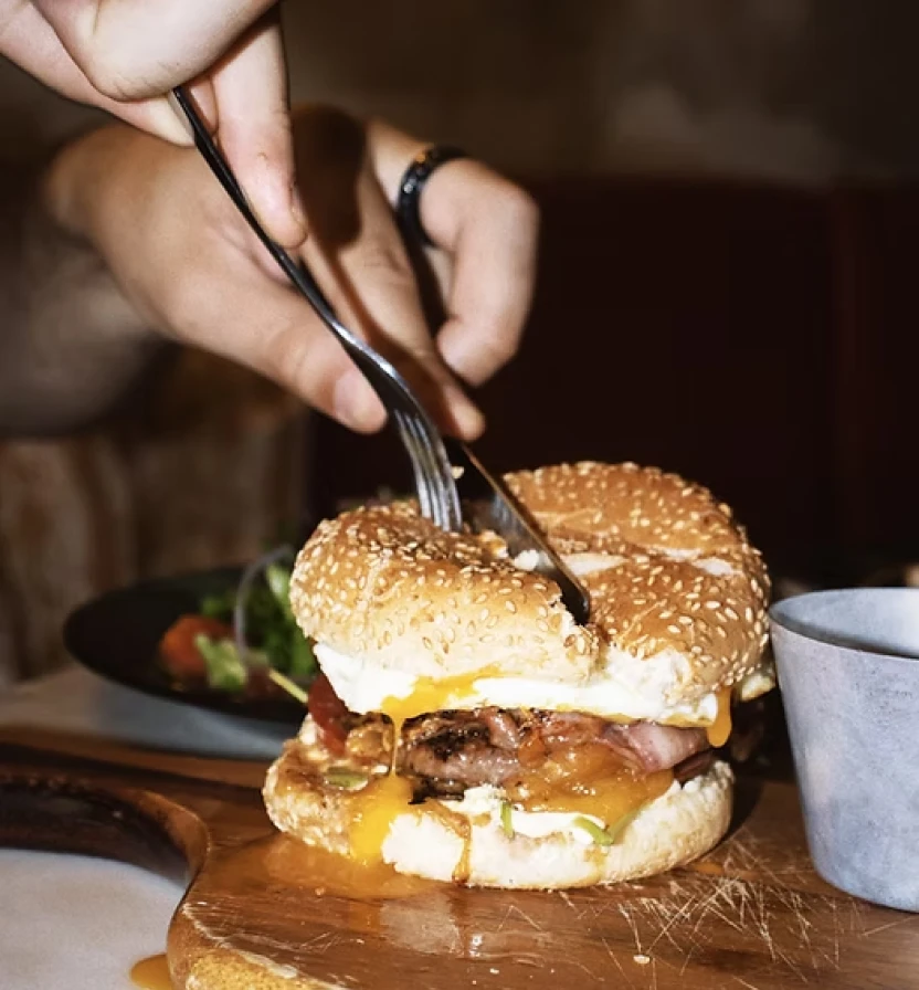 Hand slicing a burger with cheese on a wooden board, fork in view.