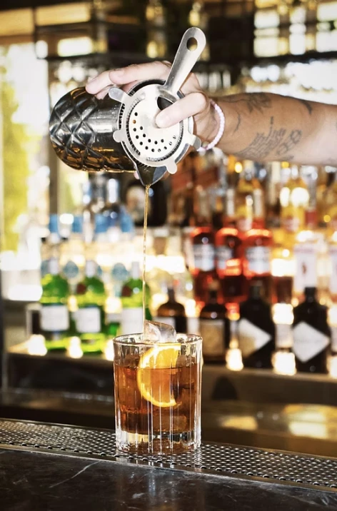 Bartender pouring amber drink from a metal jigger into a glass at bar