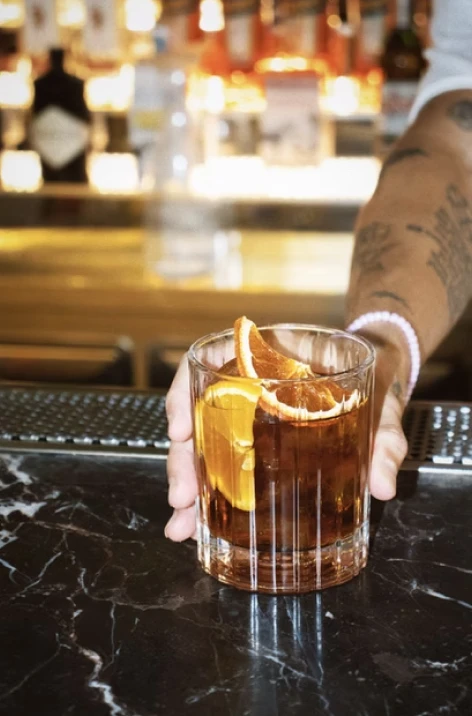 Person holding an amber cocktail with an orange garnish at a bar