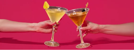 Two people clinking golden cocktails in martini glasses against pink background