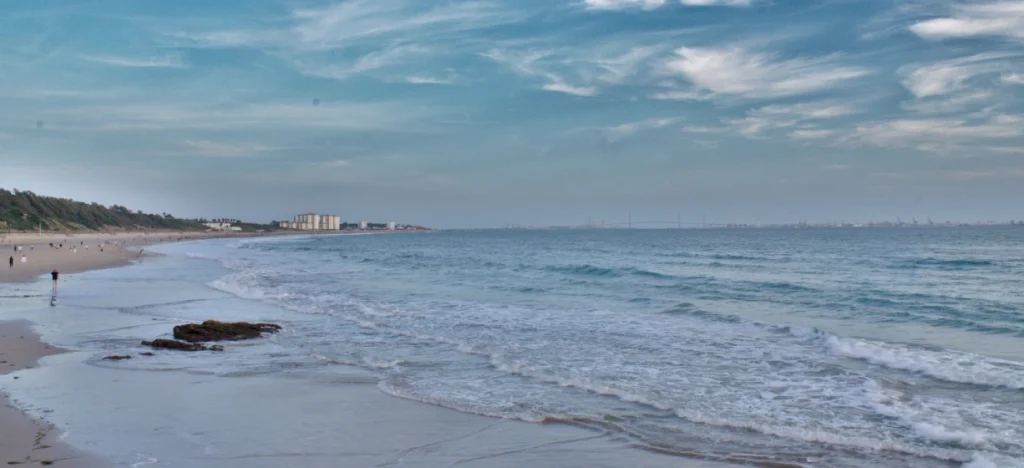 Playa extensa con olas y cielo nublado, al fondo vista costera y barcos