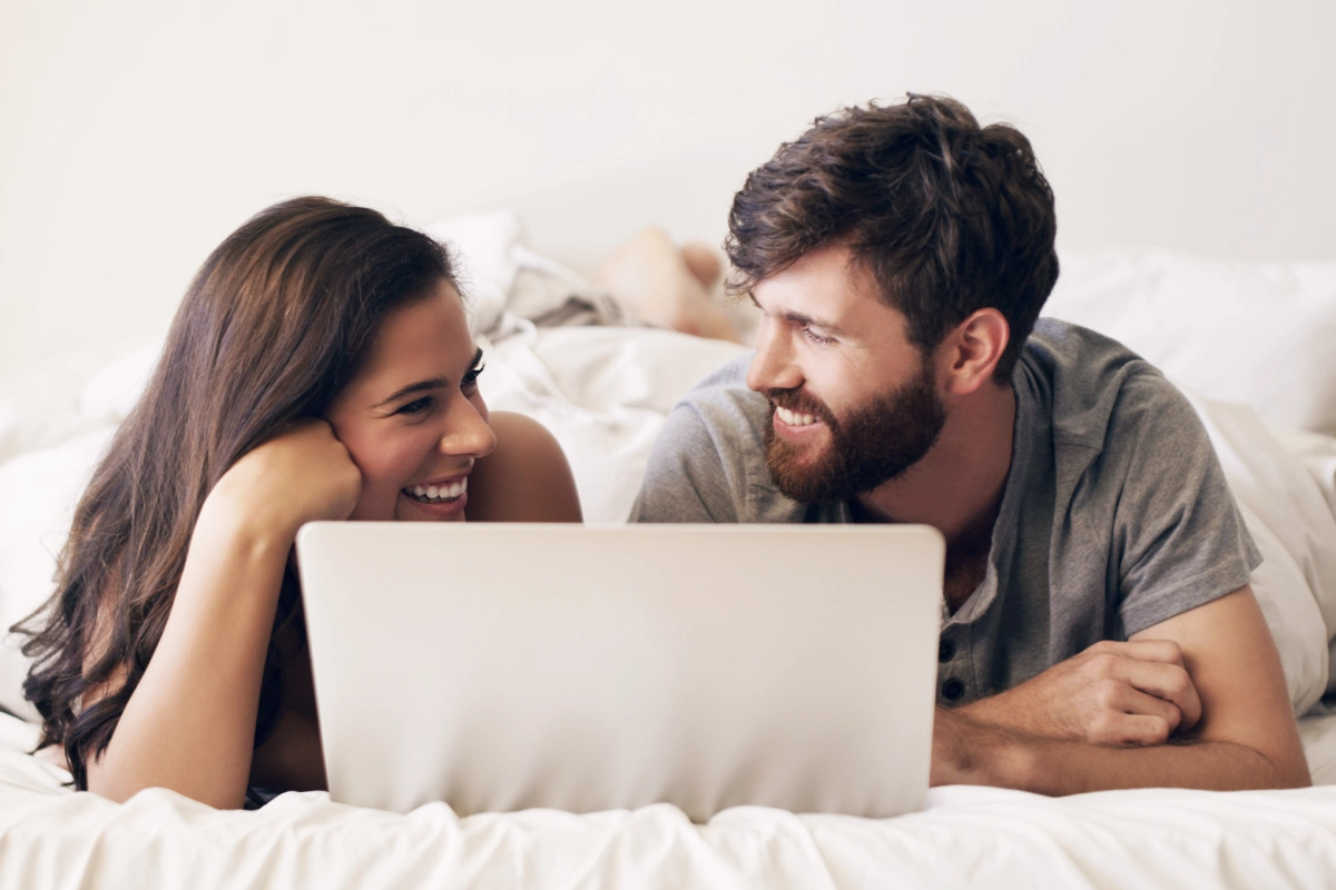 Pareja sonriendo en la cama con port&aacute;til, viendo contenido juntos