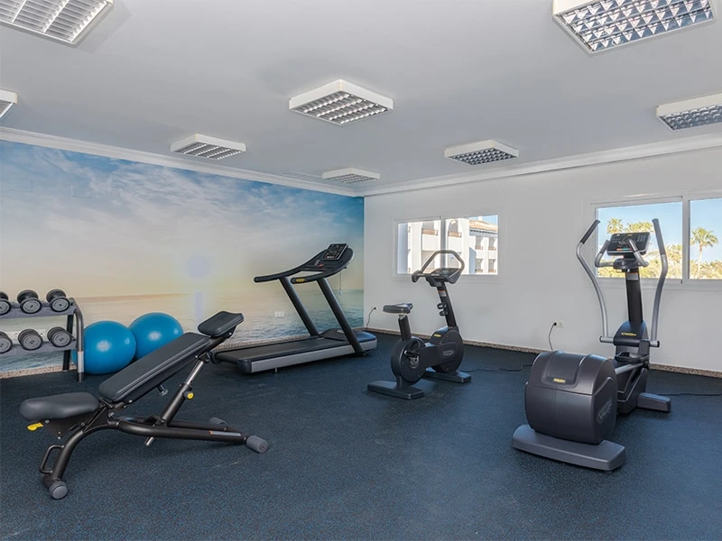 Gimnasio con cintas, bicicleta est&aacute;tica y pesas en una sala con ventanas