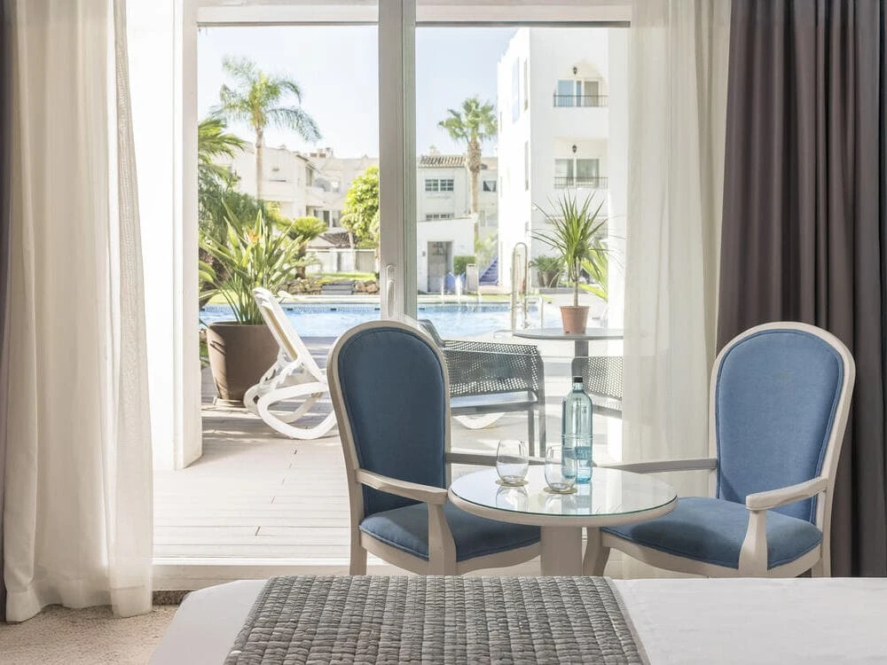 Mesa redonda con sillas azules junto a ventana y vista a piscina y palmeras