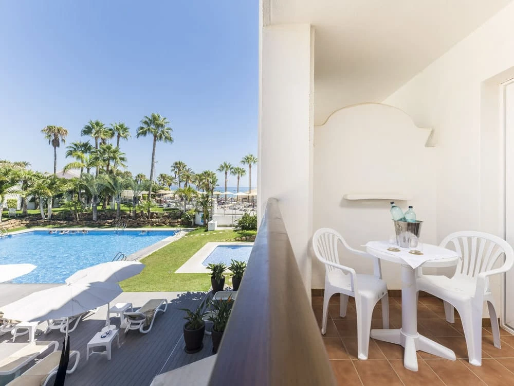 Balc&oacute;n con mesa y sillas junto a piscina, palmeras y mar al fondo