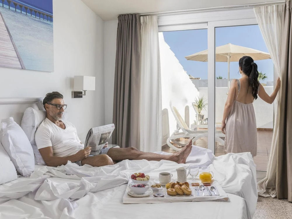 Hombre y mujer desayunan en cama junto a una ventana con vista al exterior