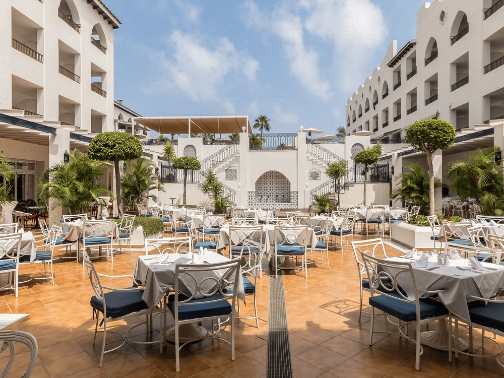 Patio de hotel con mesas y sillas, jard&iacute;n y palmeras bajo cielo despejado