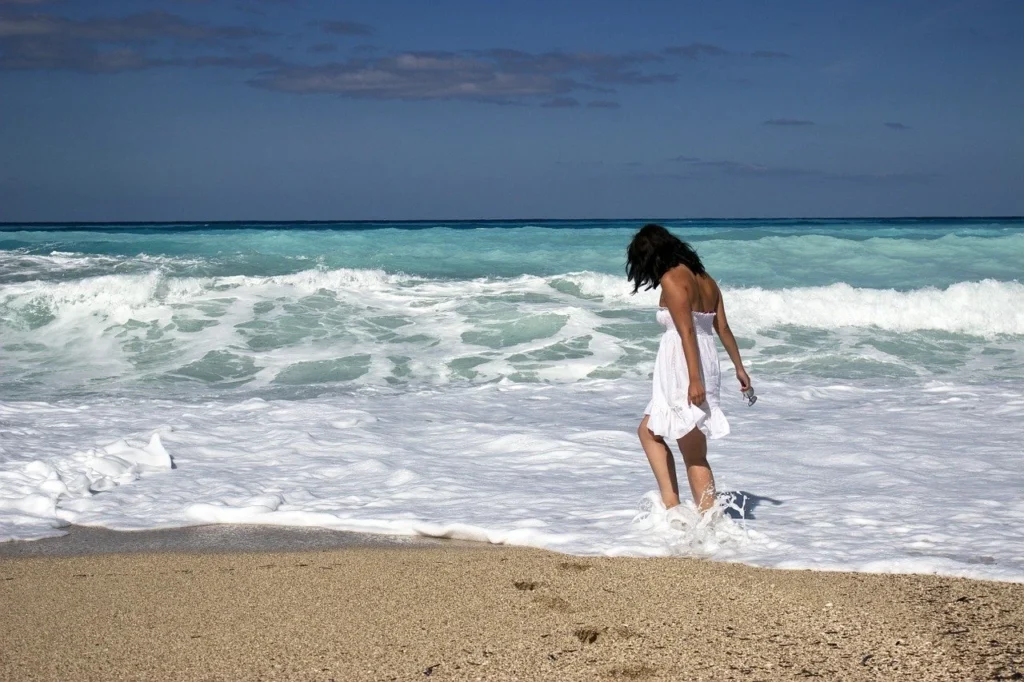 Persona caminando descalza por la orilla del mar, con olas y vestido blanco