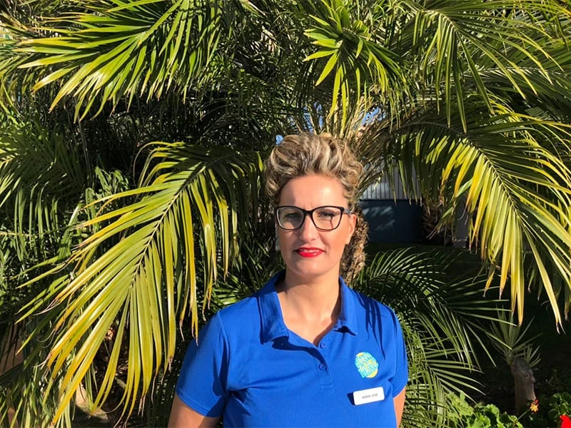 Mujer con gafas y uniforme azul frente a palmeras verdes.