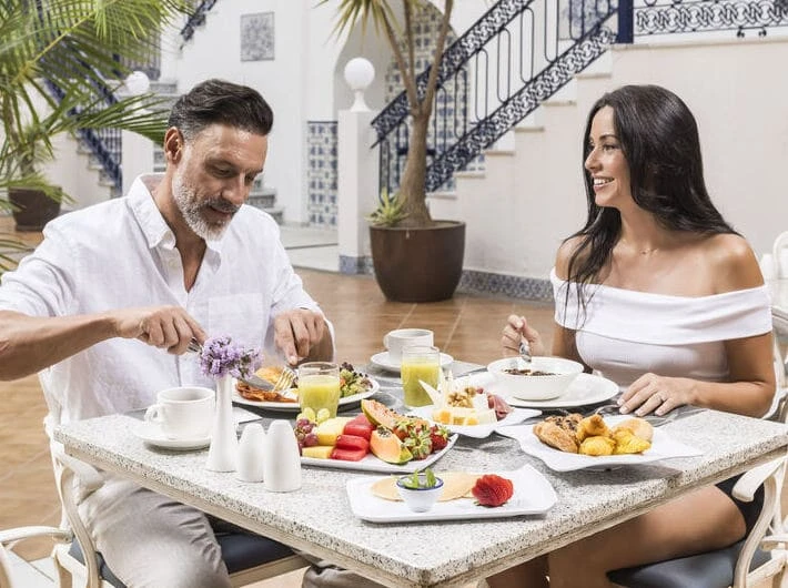 Pareja desayunando al aire libre, mesa con frutas y croissants