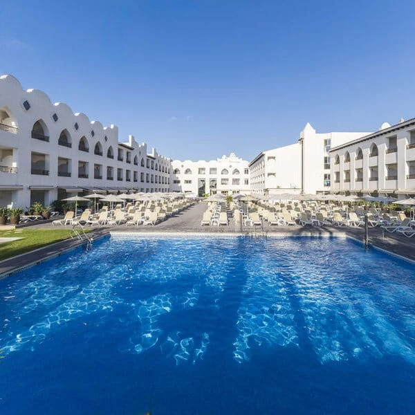 Piscina azul y tumbonas en complejo hotelero blanco, bajo cielo despejado