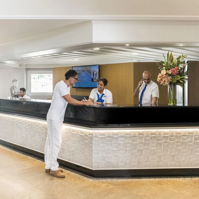 Personal de hotel atendiendo en recepci&oacute;n, con cliente y decoraci&oacute;n floral.