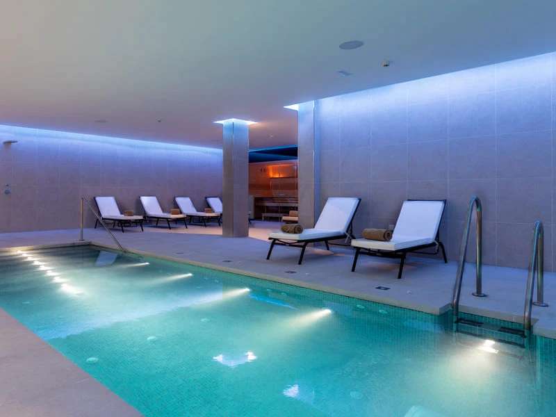 Piscina interior con tumbonas y luces c&aacute;lidas en un spa moderno.