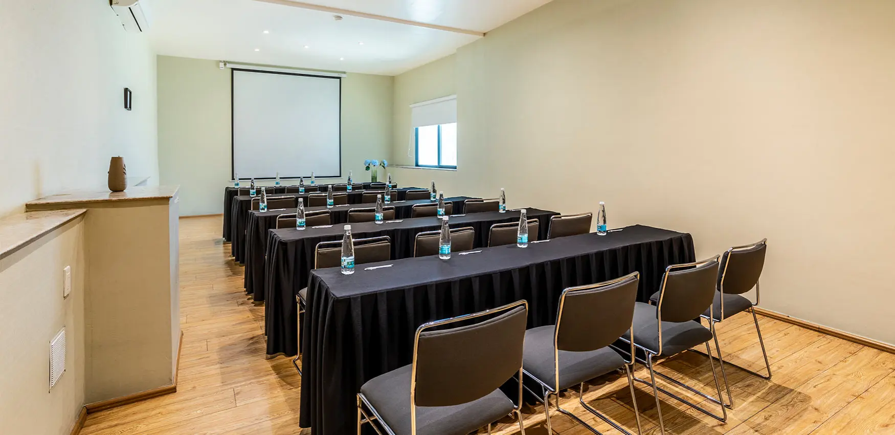 Sala de reuniones con mesas cubiertas y sillas, proyector en pantalla.