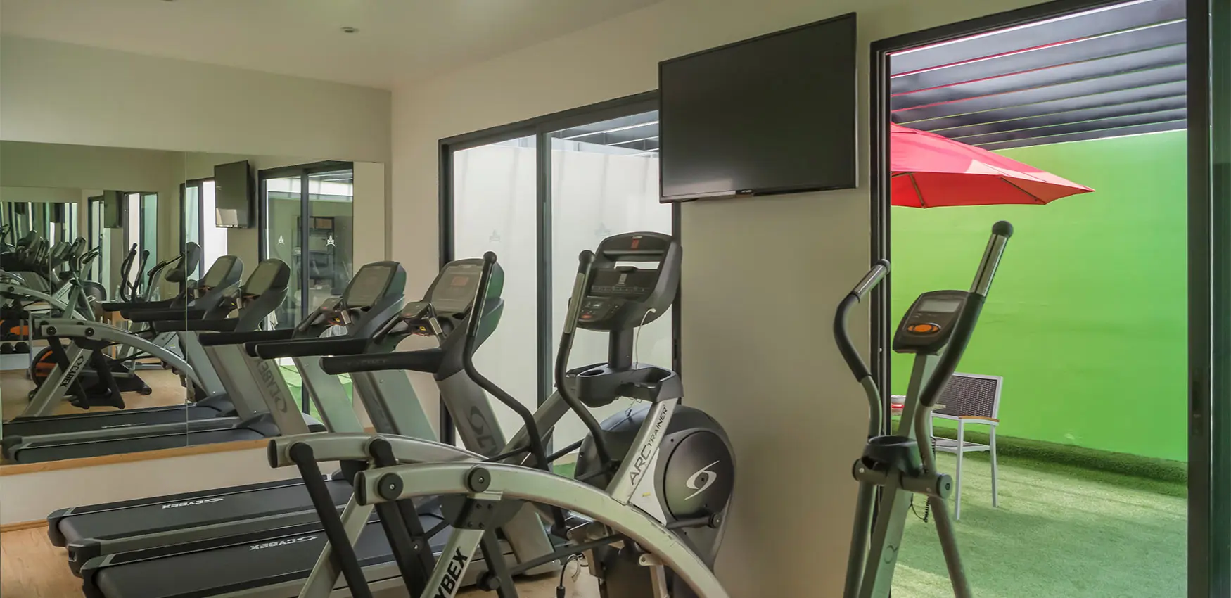 Gimnasio con máquinas de cardio y pantalla frente a fondo verde con sombrilla roja