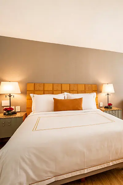 Cama con s&aacute;banas blancas y almohadas en habitaci&oacute;n de hotel, l&aacute;mparas encendidas