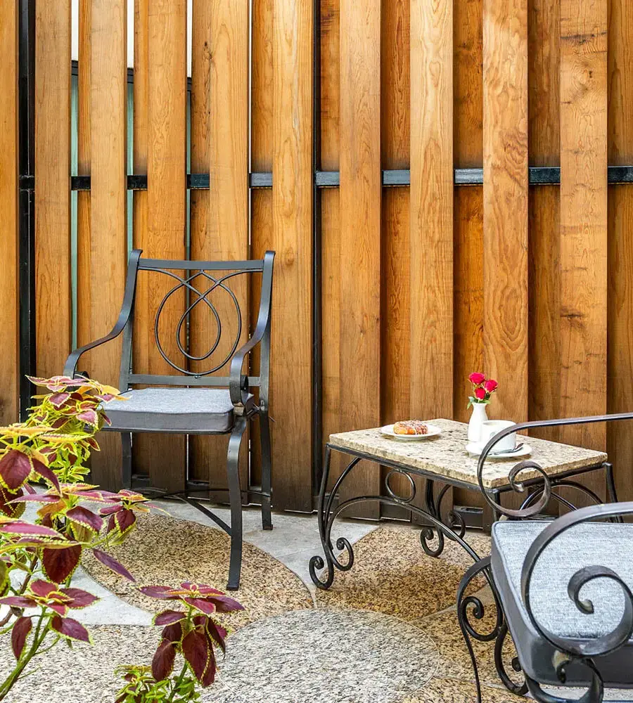 Patio con sillas met&aacute;licas, mesa de granito y flores frente a pared de madera