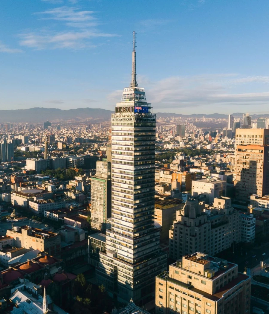 Vista aérea de un rascacielos alto en una ciudad, cielo azul y montañas al fondo.