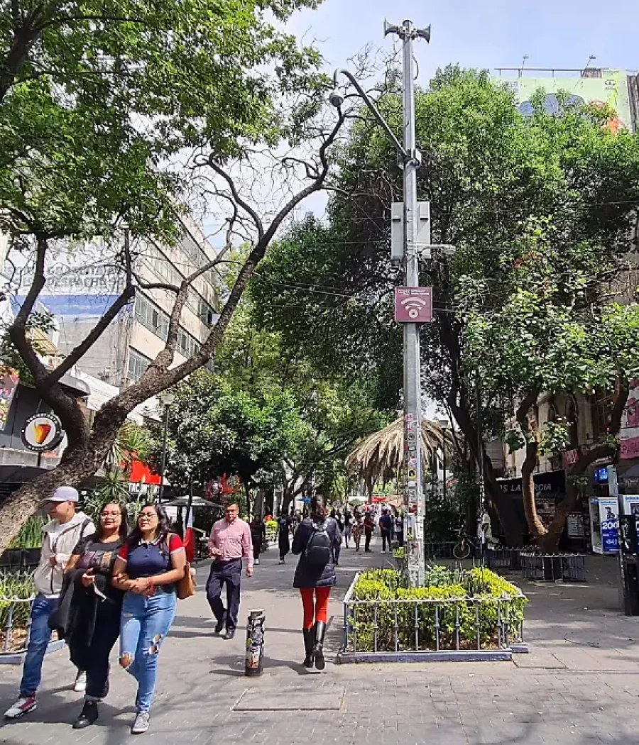Calle con peatones y un poste con señal WiFi entre árboles y edificios.
