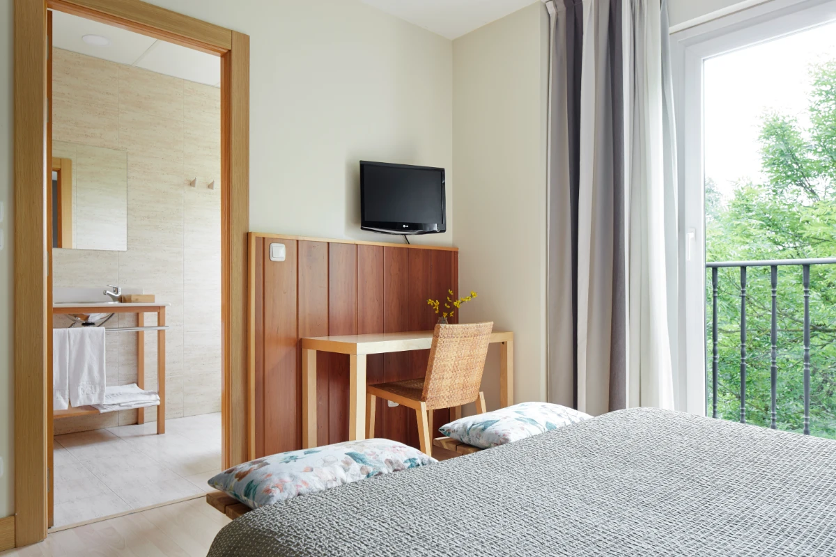 Dormitorio con cama, televisor, escritorio de madera y ventana con cortinas.