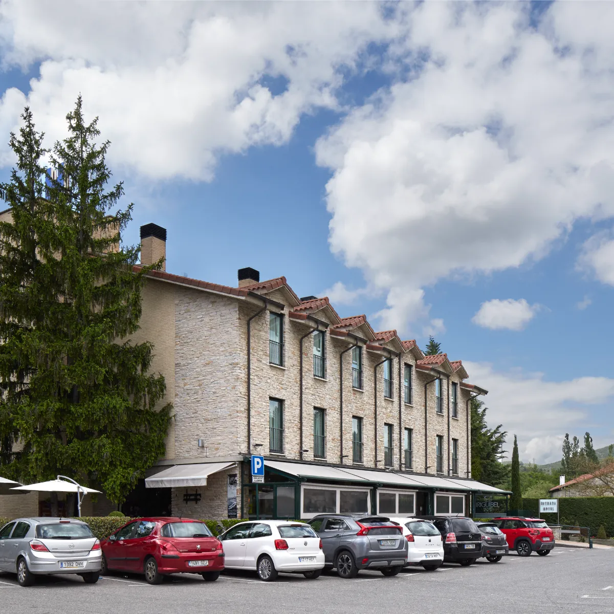 Edificio de piedra con tejado de tejas y coches estacionados bajo nubes.
