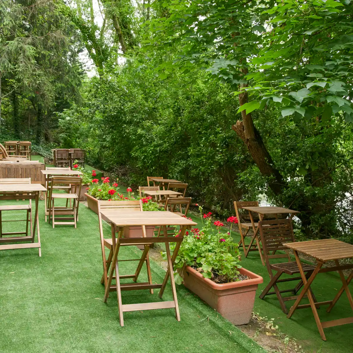 Jard&iacute;n con mesas y sillas de madera, flores rojas y vegetaci&oacute;n verde.