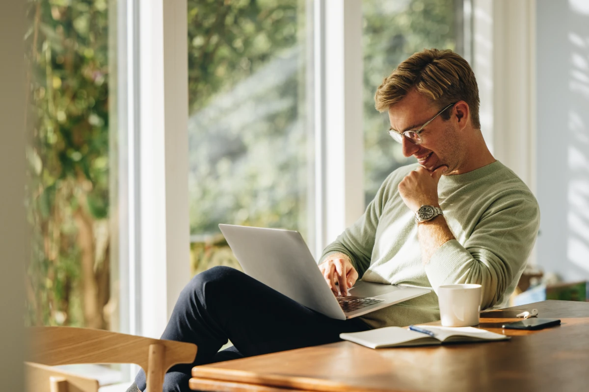 Hombre con laptop en casa, sonriendo y pensando, con taza de caf&eacute; en la mesa