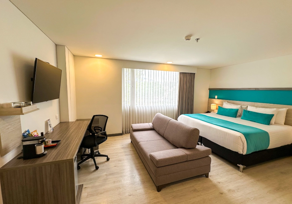 Habitaci&oacute;n de hotel con cama doble, sof&aacute; beige y escritorio con silla