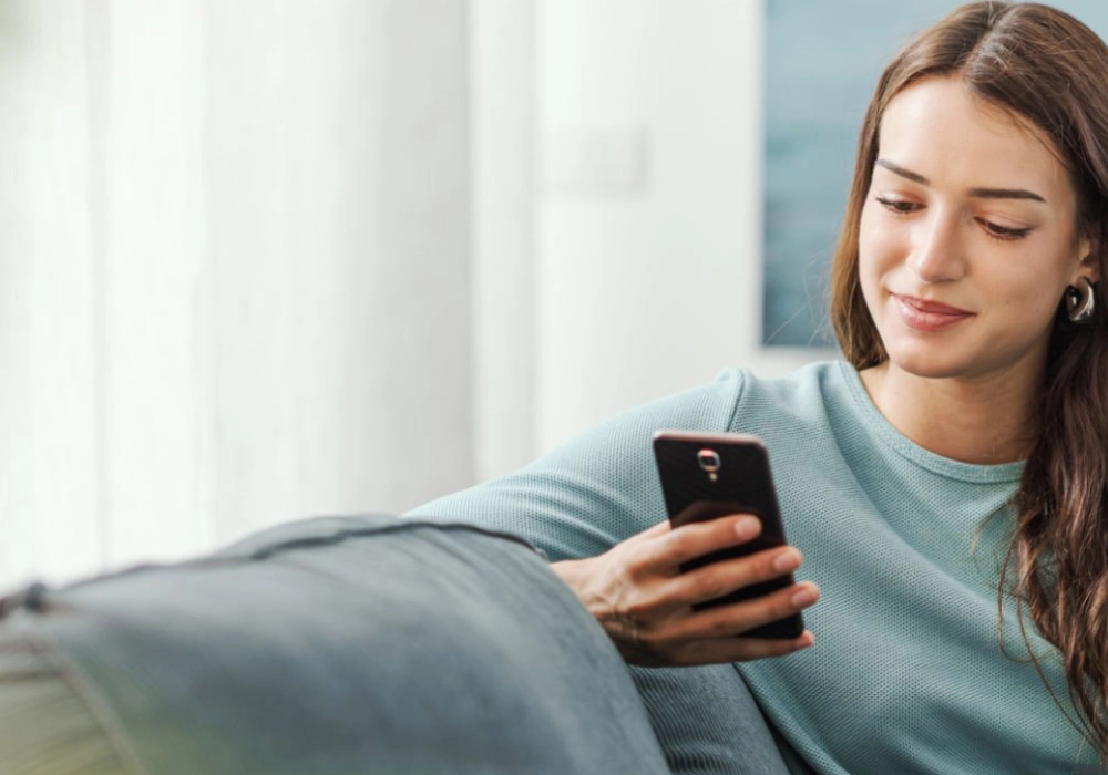 Mujer sonriente con tel&eacute;fono m&oacute;vil, sentada en un sill&oacute;n, en interior.