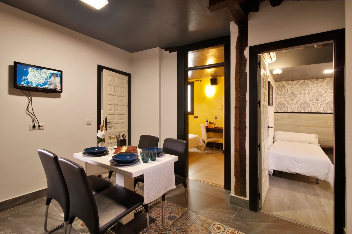 Comedor moderno con mesa y TV, y vista a dormitorio con cama y pared de ladrillo.