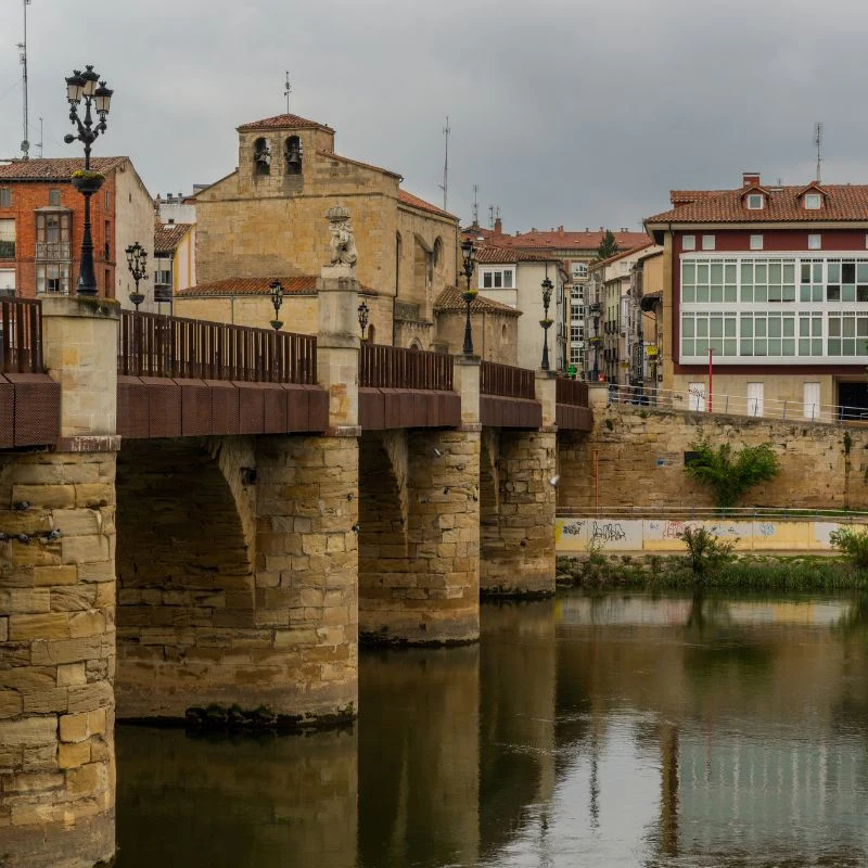 Puente romano sobre el r&iacute;o con edificios antiguos al fondo, en ciudad medieval.