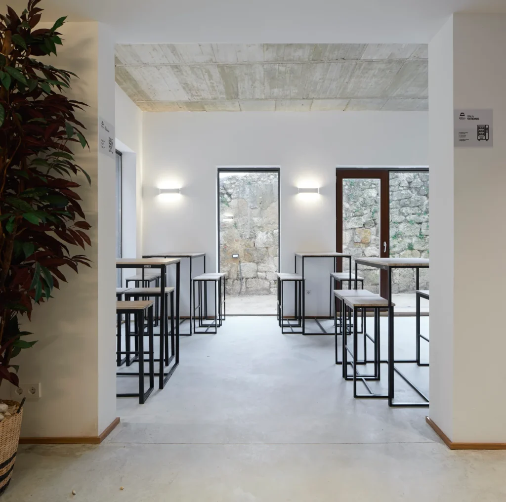 Sala tipo cafeter&iacute;a con mesas altas, paredes blancas y ventana a jard&iacute;n.