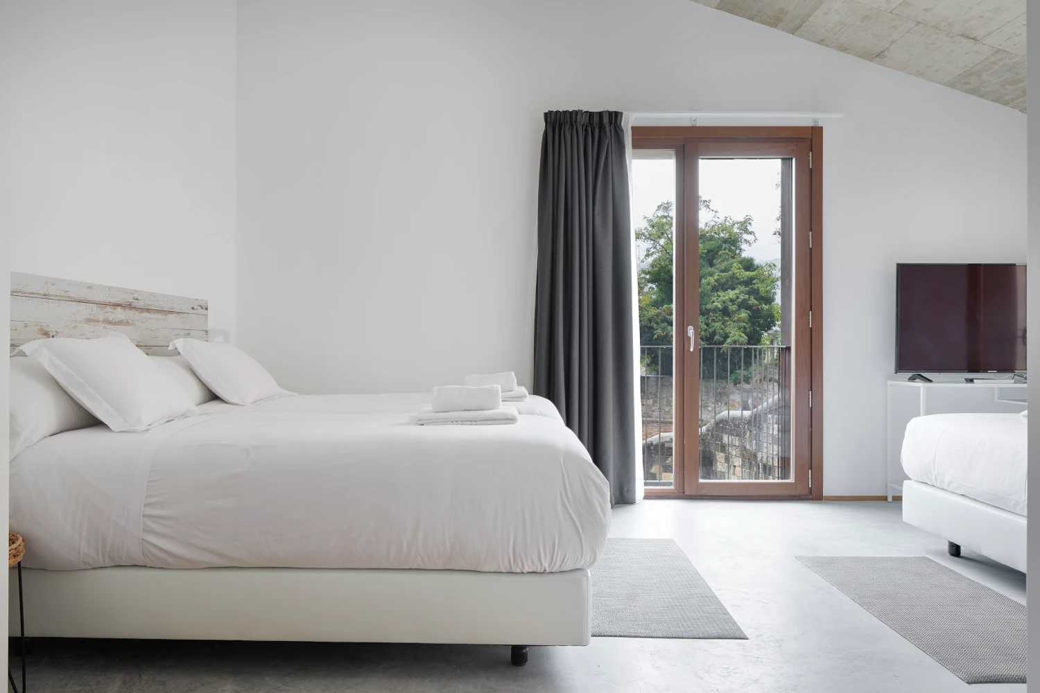 Dormitorio moderno con cama blanca, cortinas oscuras y TV junto a balc&oacute;n