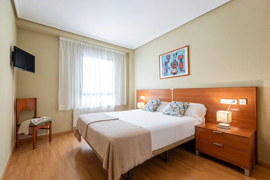 Dormitorio con cama, mesitas, l&aacute;mpara, ventana y cuadro decorativo.
