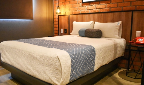 Habitaci&oacute;n de hotel con cama doble y edred&oacute;n gris sobre madera y ladrillo