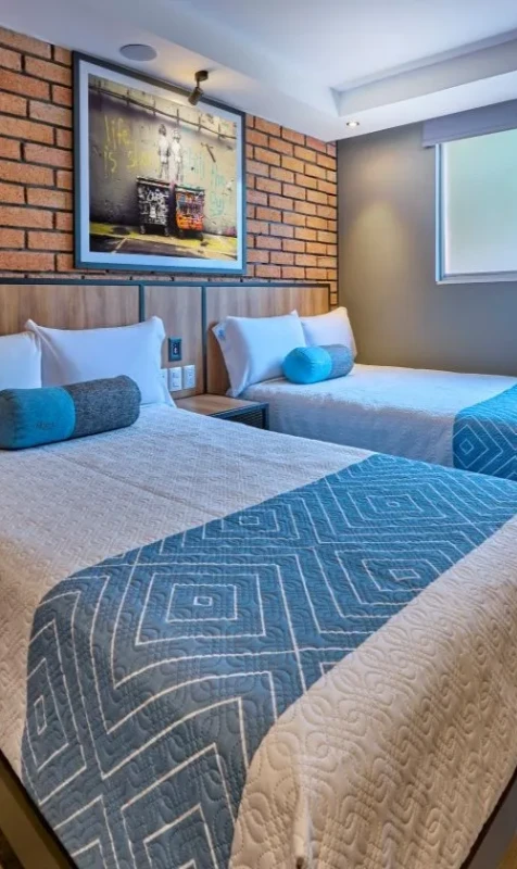 Habitaci&oacute;n con dos camas, almohadas azules, mantas y cuadro en la pared