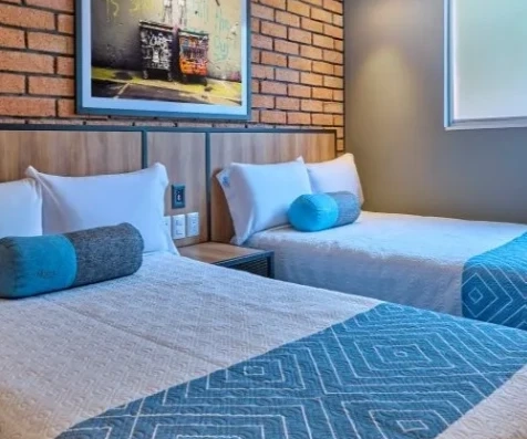 Habitaci&oacute;n hotelera con dos camas y almohadas azules sobre piso de madera