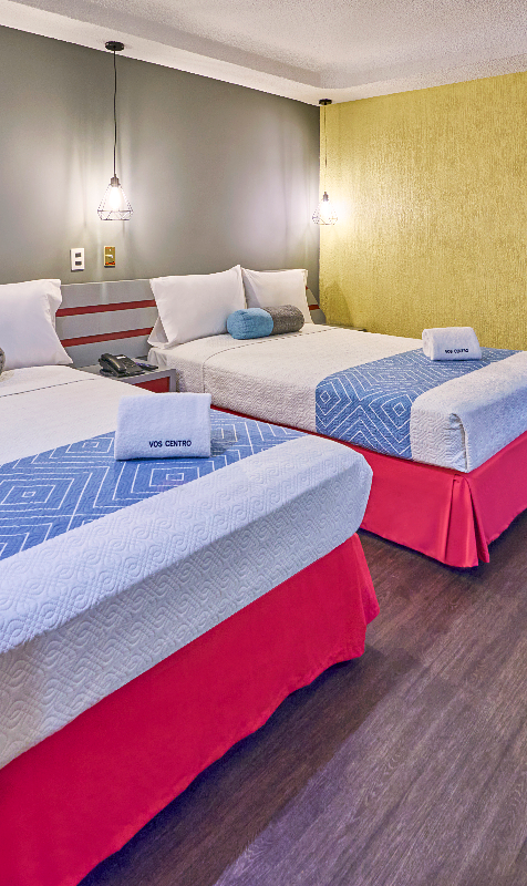 Habitaci&oacute;n de hotel con tres camas, colchas rojas y azules, y paredes grises y amarillas.