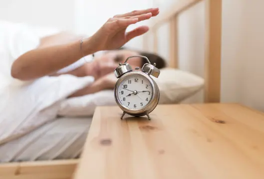 Persona estira el brazo con un despertador sobre una mesa.