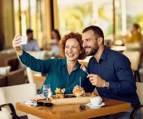 Pareja en cafeter&iacute;a tomando selfie y sonriendo, con caf&eacute; y comida en mesa