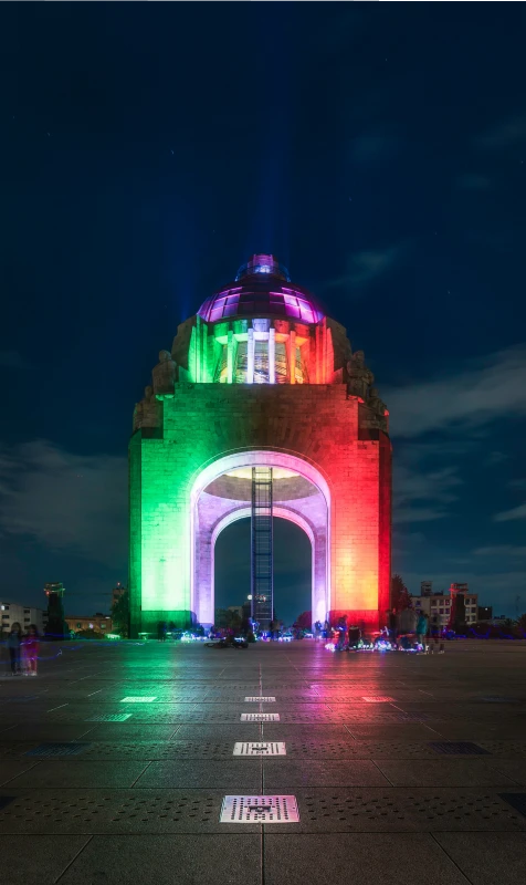 Torre iluminada con luces de colores sobre el pavimento de una plaza nocturna.