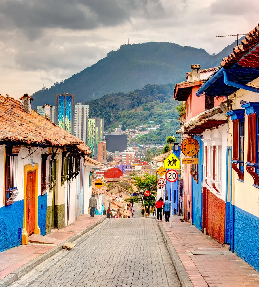 Calle colonial con casas pintadas y monta&ntilde;as al fondo, con peatones.