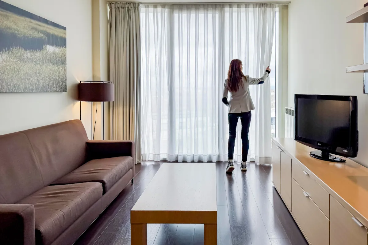 Persona frente a cortinas blancas en sala con sofá, TV y lámpara de pie.