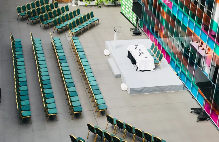 Arreglo de sillas verdes y mesas blancas en un auditorio con telones de colores.