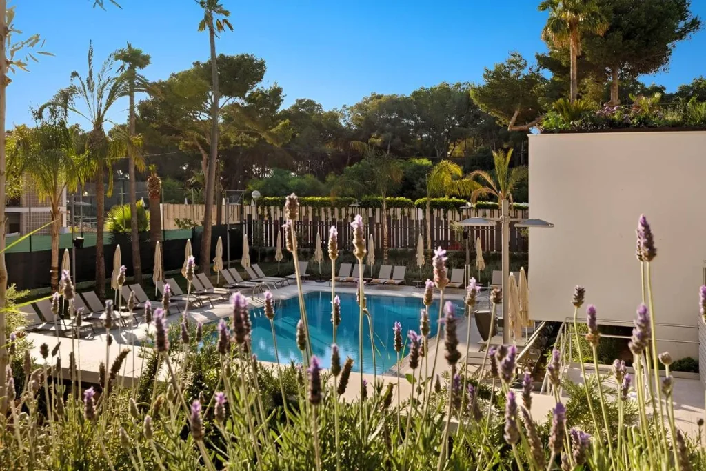 Piscina al aire libre con palmeras, flores moradas y tumbonas en hotel.